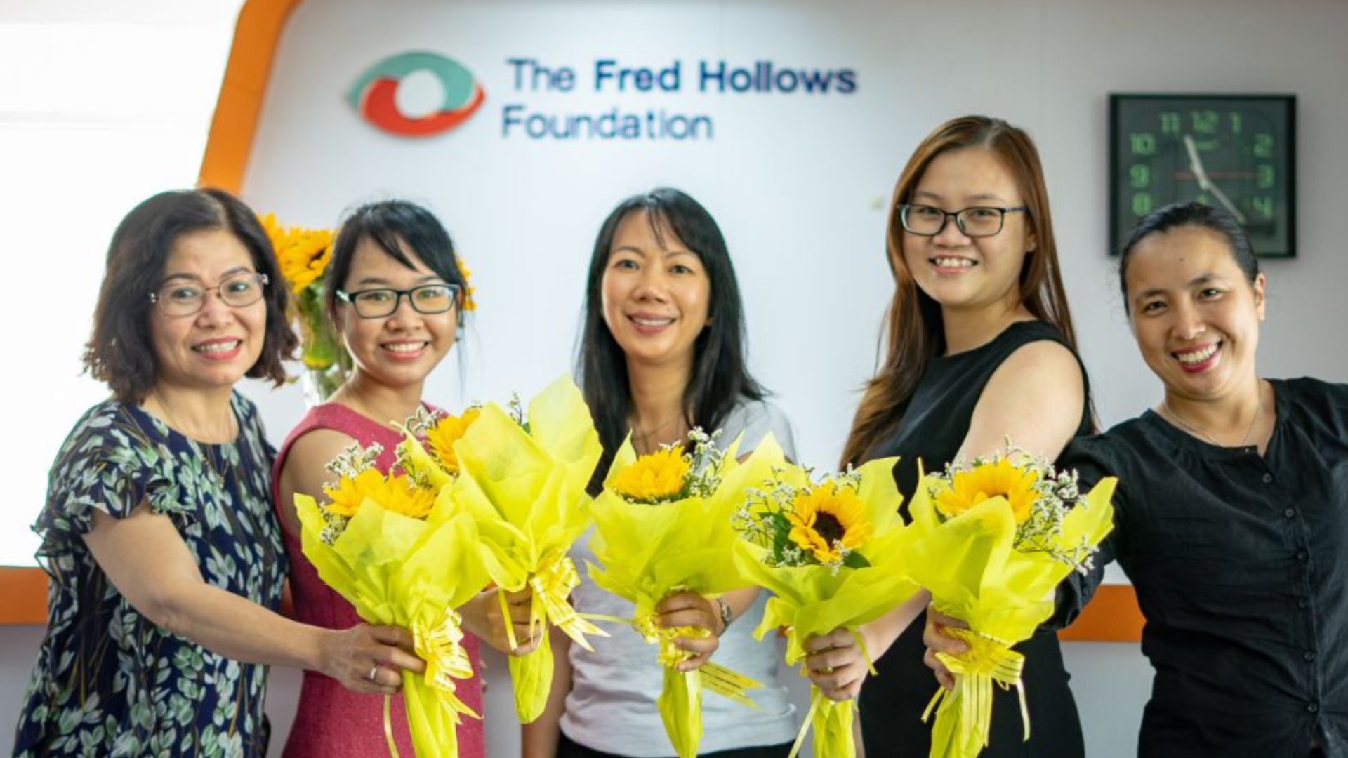 Four people standing next to each other holding yellow flowers standing in front of a Fred Hollows Foundation sign