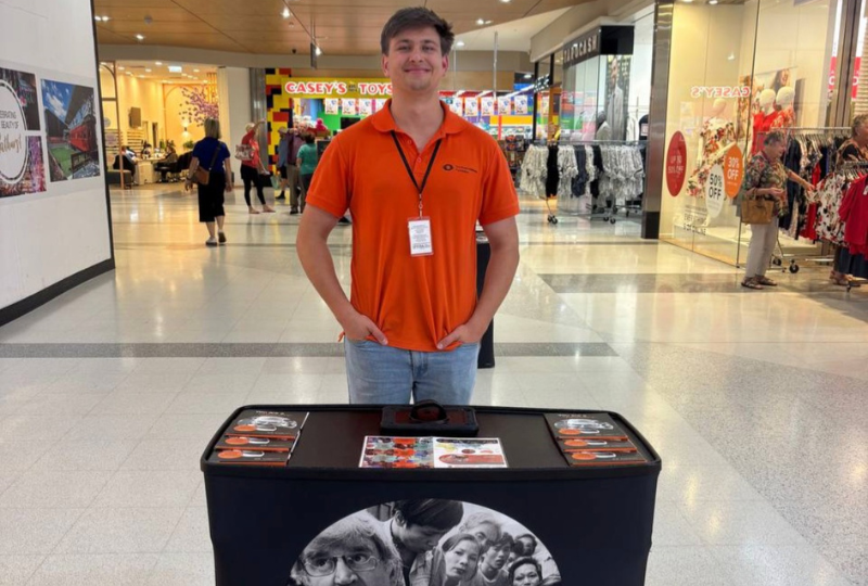 A man wearing an orange Fred Hollows Foundation polo. He is standing behind a table with information packs about The Foundation.