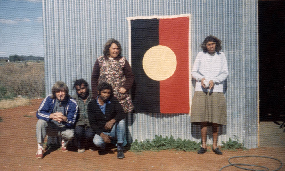 Fred Hollows crouching down next to a group of Indigenous people in front of the Aboriginal flag