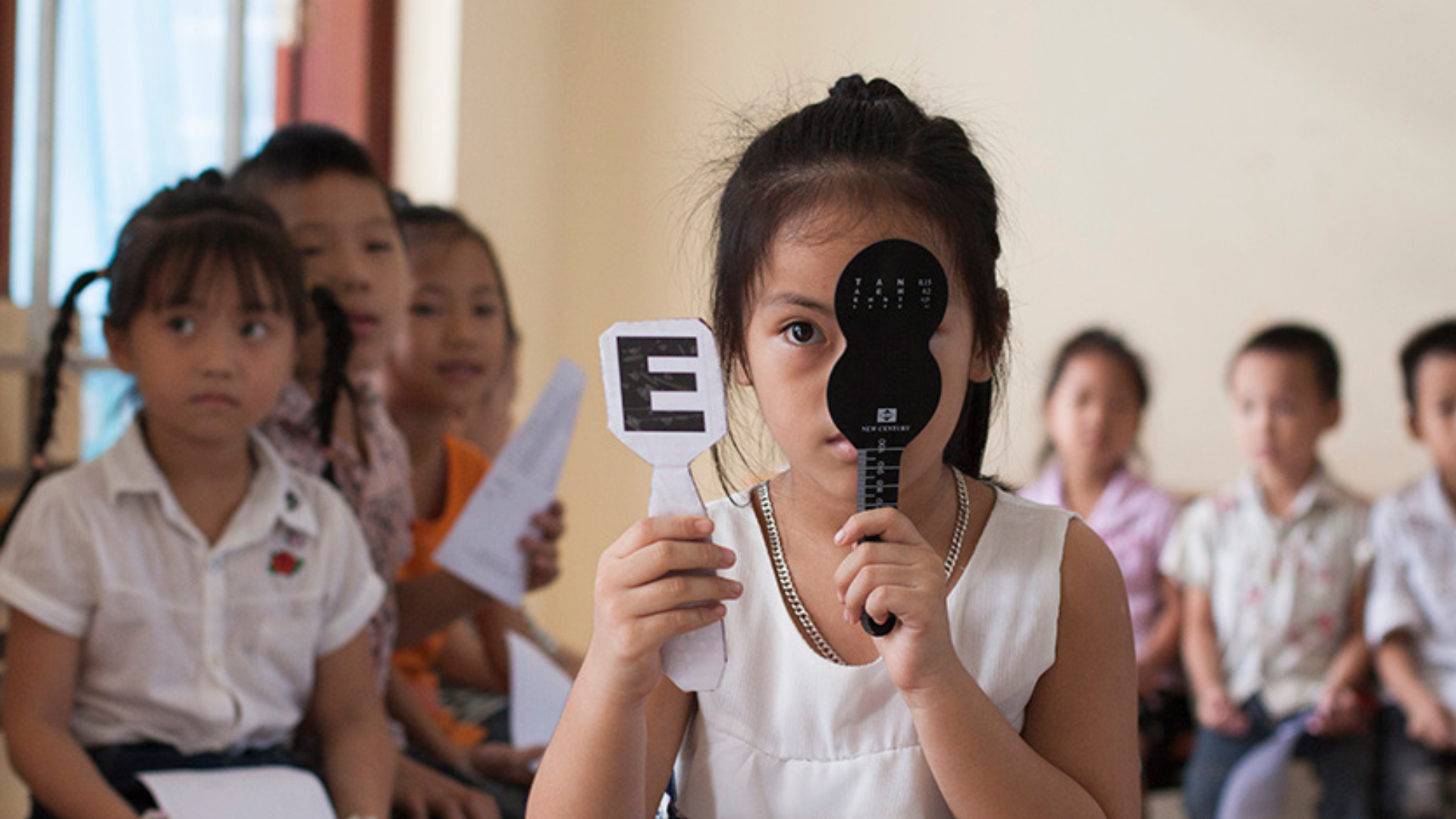 A Vietnamese girl having her eyes screened. She is holding up a card with the letter 'E' on it in one hand, and blocking her right eye with the other hand.