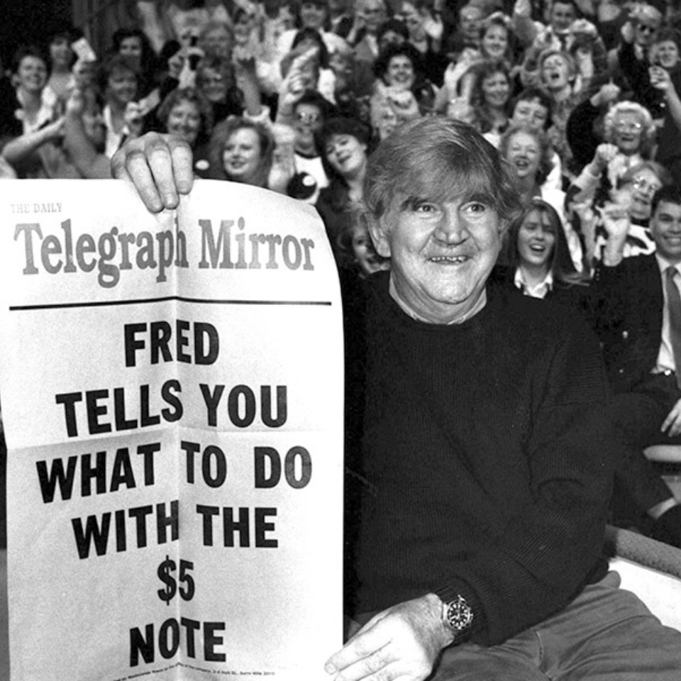 Fred on a TV show with a crowd of people in the background. He is holding up a print that says 'Fred tells you what to do with the $5 note'