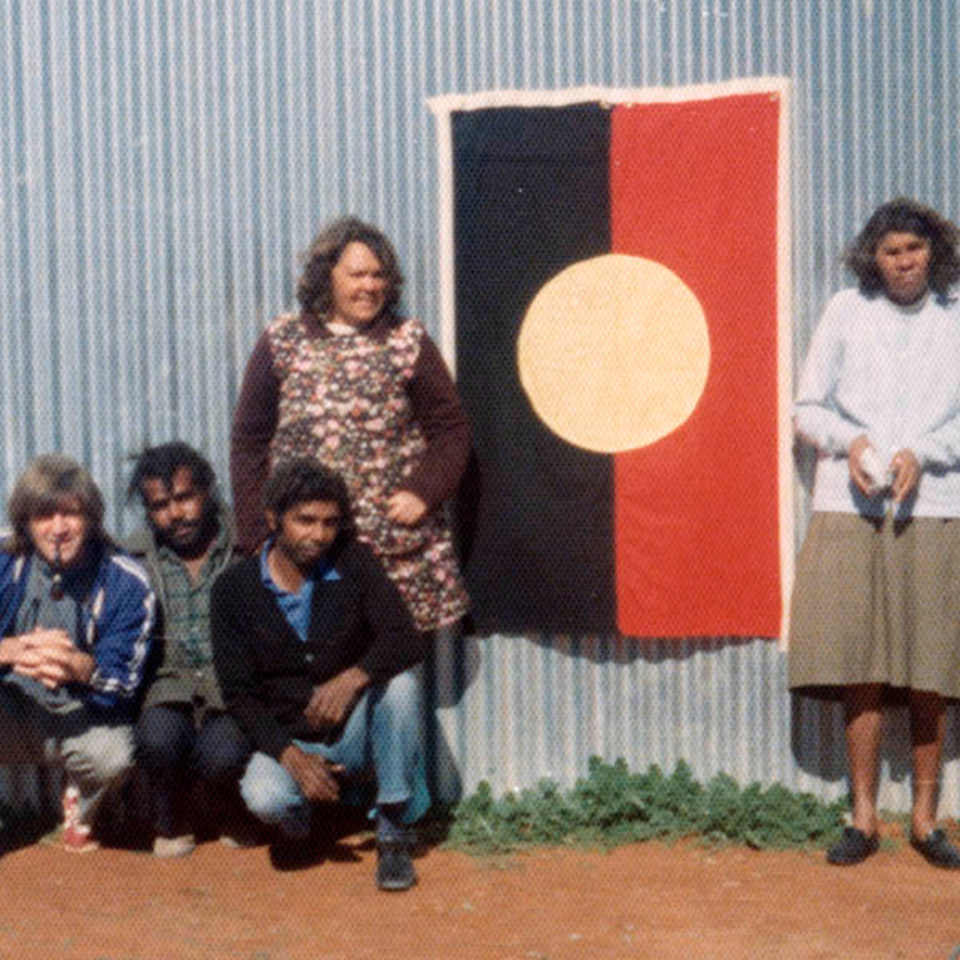 Fred crouched down posing with an indigenous group in front of the Aboriginal flag