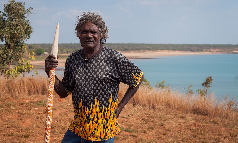 Aboriginal man holding an engraved spear with a body of water in the background