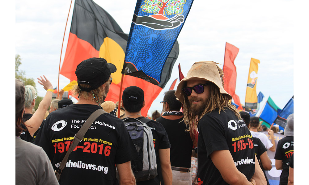 Man in foreground wearing National Trachoma Eye Health shirt, looking back at camera with Indigenous and Torres Strait flags in the background during Freedom Walk