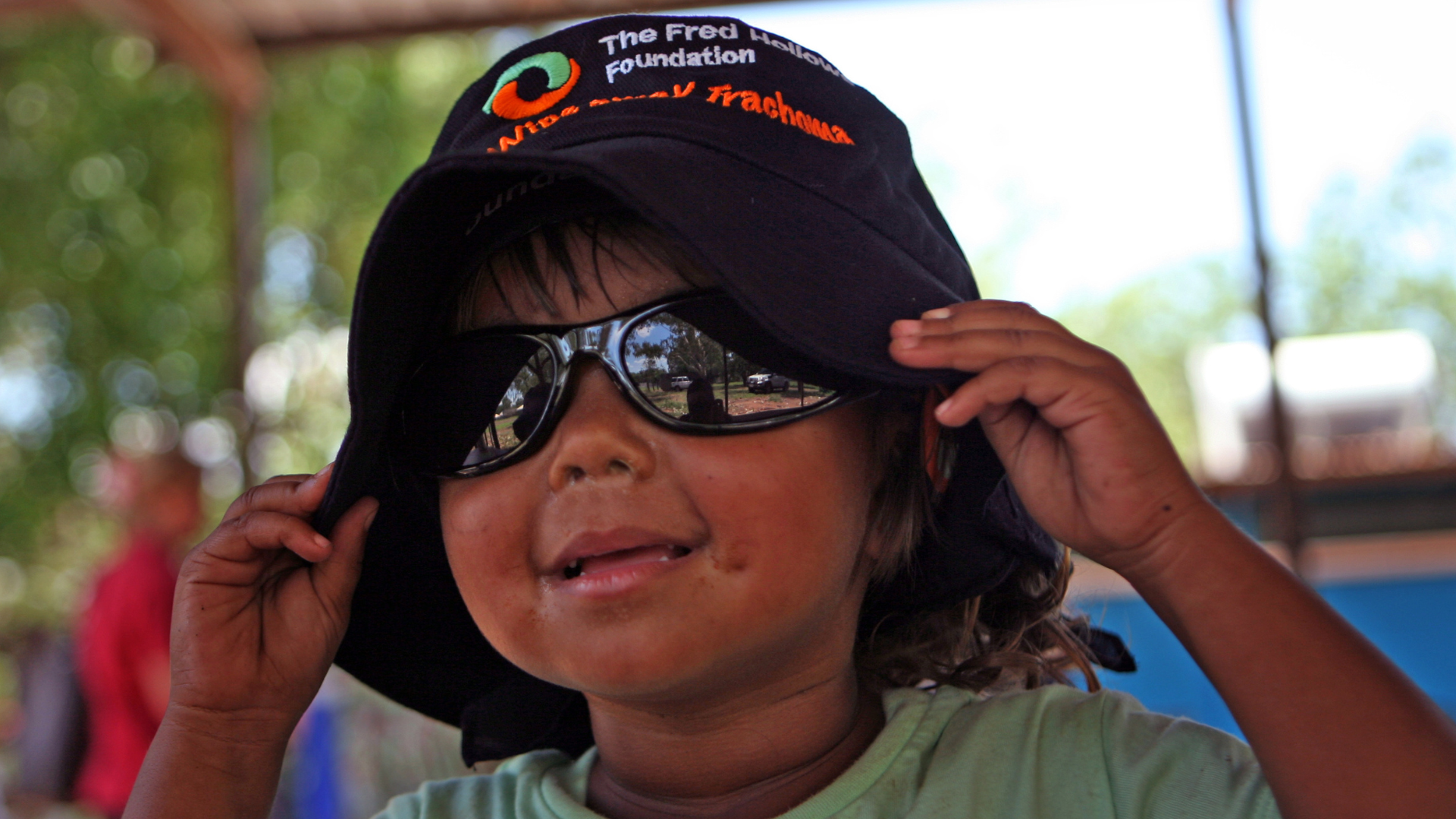 A young indigenous boy wearing sunglasses and a Fred Hollows Foundation hat smiling at the camera