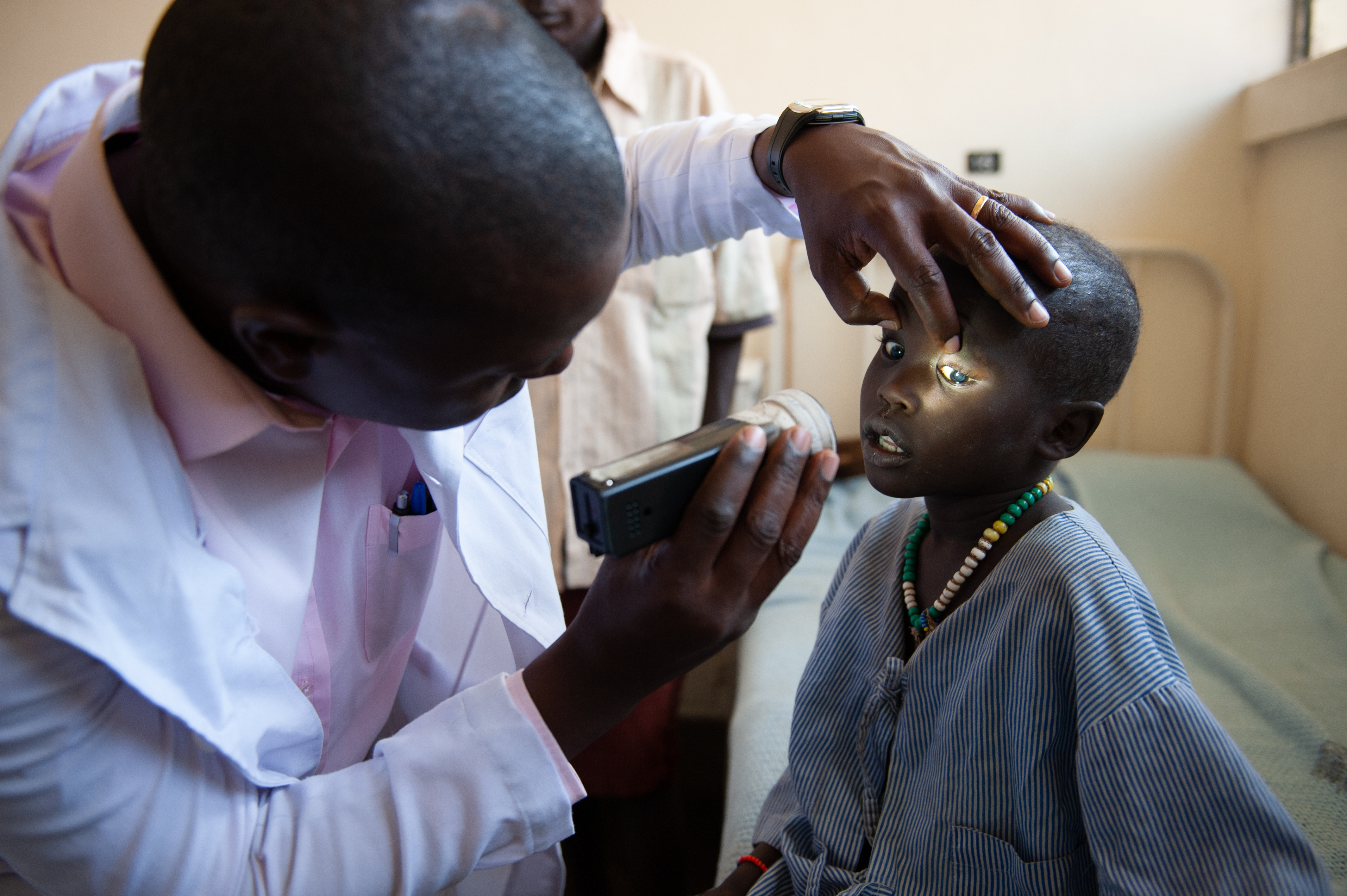 Kipar sitting on a hospital bed as the Dr examines his eyes. Kipar's father is standing in the background watching on