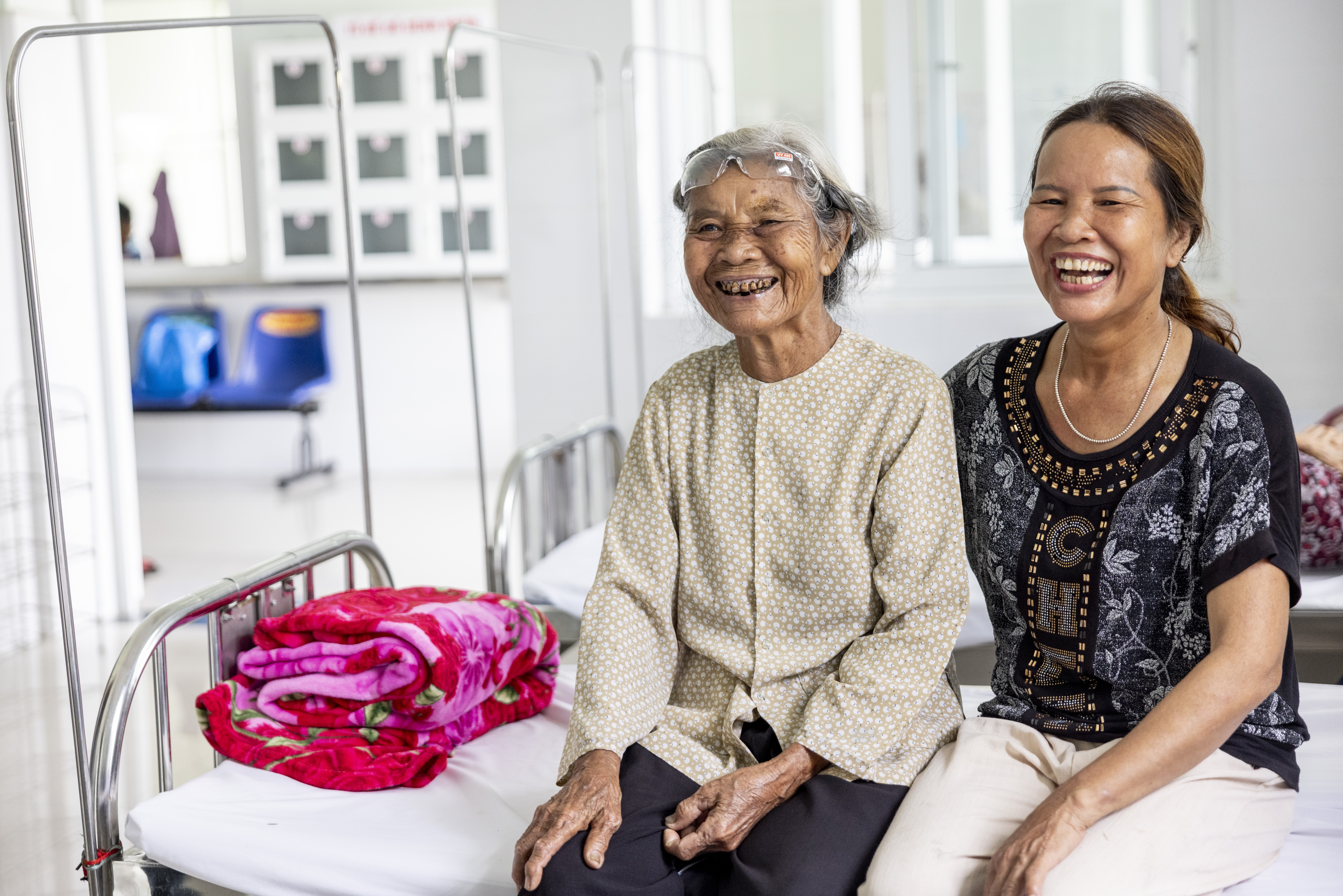 Den sitting on a hospital bed with her daughter in law after having her eye patch removed. They are both smiling wide and laughing